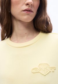 Κοντινό πλάνο γυναίκας που φοράει ανοιχτό κίτρινο φούτερ με λ ογότυπο GANT στο στήθος, εμφανίζονται μόνο τα χείλη, ο λαιμός και μέρος από τα καστανά μαλλιά της.