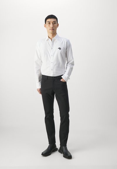 Chemise blanche à manches longues avec un petit logo noir, associée à un pantalon noir slim-fit et des bottines noires.