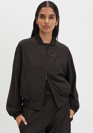 Frau mit langen dunklen Haaren, die eine schwarze Bomberjacke mit Reißverschluss und schwarze Hose trägt, steht mit den Händen in den Taschen vor einem einfarbigen Hintergrund.