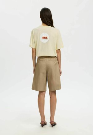 Femme aux longs cheveux foncés portant un t-shirt jaune clair avec un motif d'assiette de nourriture dans le dos, un short beige à hauteur des genoux et des talons marron.