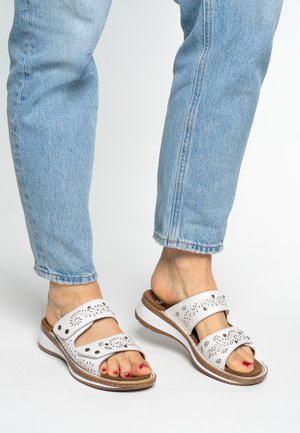 Weiße Ledersandalen mit zwei perforierten Riemen, braunem Boden und weißer Sohle. Kombiniert mit hellblauen, gekürzten Jeans, die die Zehen zeigen.
