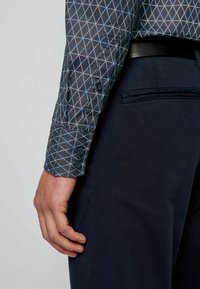 Donkerblauwe shirt met een geometrisch lichtblauw patroon, met knoopmanchetten, gecombineerd met marineblauwe broeken, waarbij een deel van een hand op de tailleband zichtbaar is.