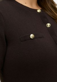 Tumepruuni soonikkoes kardigan ümara kaelusega, millel on kuldsed nupud ja tekstuuriga paneel taskuga. Sile kangas ja keha järgnev lõige.