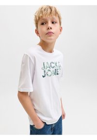 Ξανθός αγόρι με λευκό μπλουζάκι "Jack & Jones" και μπλε τζιν, στέκεται με το ένα χέρι στην τσέπη, κοιτάζοντας προς τα πλαϊνά, μπροστά σε λευκό φόντο.