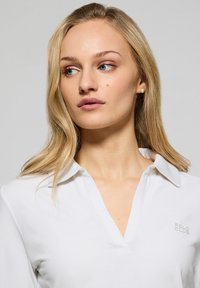 Vit långärmad pikétröja med V-ringning och vikt krage. Har en diskret "POLO CLUB"-logotyp på vänster sida. Slät tygtextur.