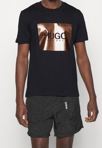 Schwarzes Baumwoll-T-Shirt mit einem glänzenden bronzefarbenen rechteckigen Logo mit dem Text "HUGO" und "HUGO BOSS". Kombiniert mit schwarzen strukturierten Shorts.