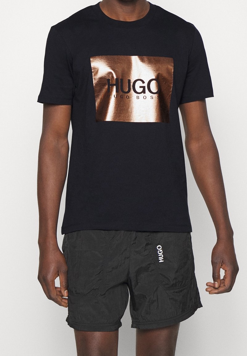 Schwarzes Baumwoll-T-Shirt mit einem glänzenden bronzefarbenen rechteckigen Logo mit dem Text "HUGO" und "HUGO BOSS". Kombiniert mit schwarzen strukturierten Shorts.