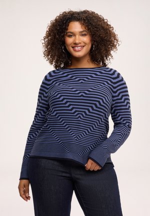 Donna sorridente con capelli ricci che indossa una maglia a maniche lunghe a righe blu e nere e jeans scuri su uno sfondo chiaro uniforme.