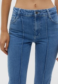 Jean en denim bleu à taille haute, doté d'une fermeture éclair, de poches avant et de surpiqûres contrastantes le long des coutures latérales.