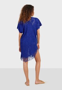 Kaftán azul de encaje con mangas cortas, que presenta un dobladillo con flecos y un patrón floral, mostrando una textura transparente para una cobertura ligera.