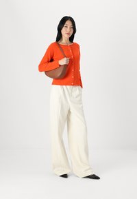 Orange knitted cardigan with buttons, wide white trousers, and a brown curved handbag. Model stands against a neutral background.