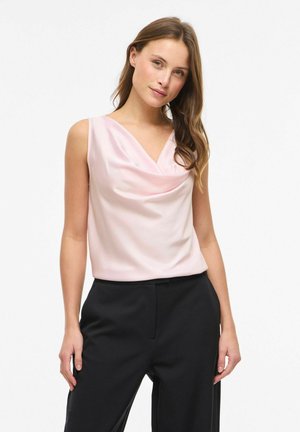 Donna con capelli castano lunghi che indossa una canotta rosa pallido senza maniche con collo a scialle e pantaloni neri a vita alta, in piedi contro uno sfondo bianco.