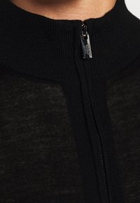 Černý pletený svršek na zip s vysokým límcem, který má hladkou texturu a kovový zip u krku.