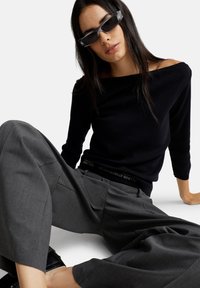 Top de punto negro con hombros al descubierto, combinado con pantalones de pierna ancha grises de tejido texturizado y una cinturilla con logo visible. Incluye gafas de sol grises.