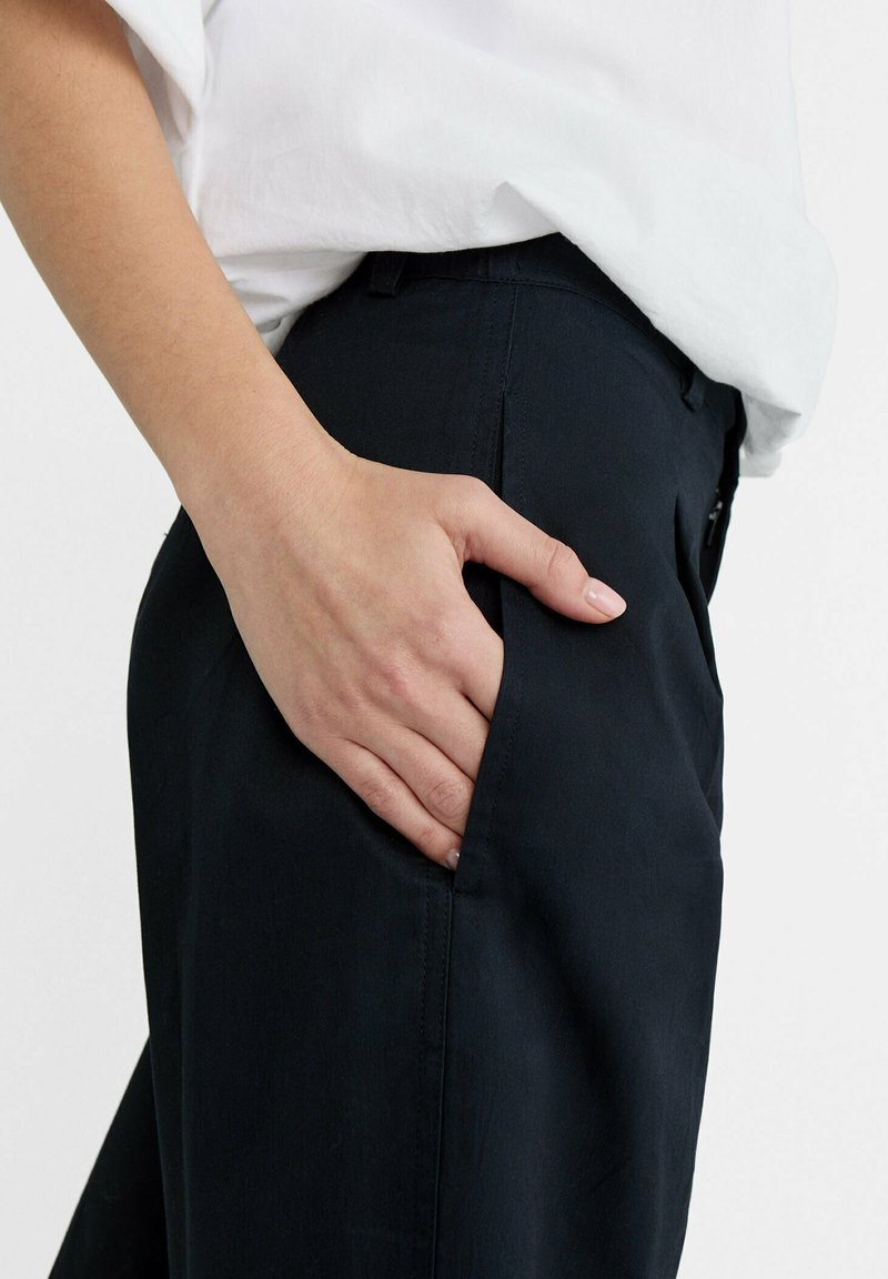 Pantaloni navy realizzati in tessuto liscio, dotati di una tasca laterale, vestibilità su misura e un dettaglio di cucitura sottile vicino alla vita. La maglietta è bianca.