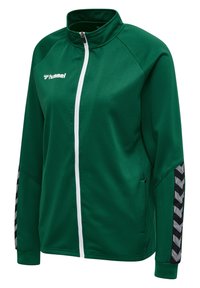 Groene ritssluitingjack gemaakt van gestructureerde stof, met een witte rits, vo pockets en zwarte chevron-accenten op de mouwen.