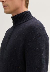 Marineblauwe gebreide trui met een hoge geribbelde kraag en een halve rits, voorzien van veelkleurige spikkels en een logopatch op de mouw.