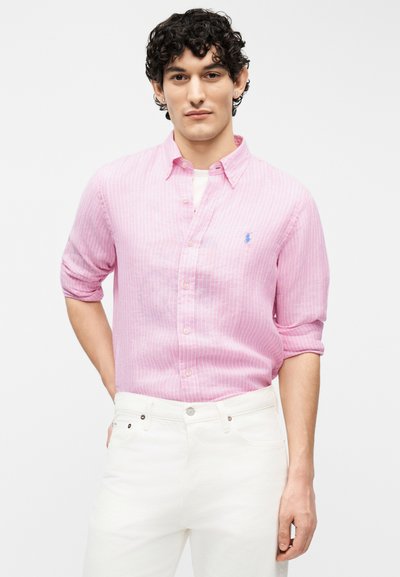 Άνδρας που φοράει ανοιχτό ροζ πουκάμισο με ρίγες και κουμπιά, με τα μανίκια σηκωμένα, και άσπρο παντελόνι, στέκεται μπροστά σε απλό φόντο.