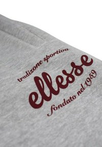 Tissu gris à texture douce présentant une broderie bordeaux avec l'inscription "ellesse" ainsi que les petits textes "tradizione sportiva" et "fondato nel 1959."