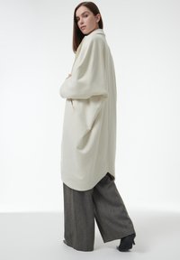 Abrigo crema de gran tamaño con un collar doblado, aberturas laterales y textura suave, combinado con pantalones de pierna ancha grises y tacones negros.