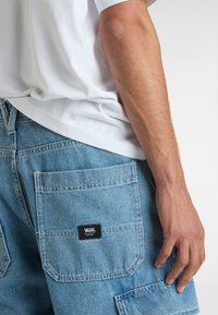 Pantaloni corti in denim blu chiaro con più tasche, dettagli cuciti e un'etichetta con il logo Vans nera. Una maglietta bianca scende sulla vita.