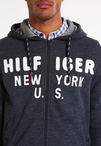 Felpa con zip di colore navy con scritta bianca "HILFIGER NEW YORK U.S." e cappuccio interno grigio. Presenta un tessuto testurizzato e cordini con motivo.