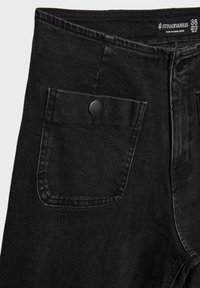 Jean en denim noir en gros plan montrant une poche avant boutonnée, une ceinture avec une étiquette indiquant la taille 38 et la marque Stradivarius.