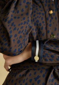 Brūna auduma ar tumši zilu leopardu rakstu, ar zelta pogām un svītrainu piedurkni zilā, baltā un melnā krāsā. Mīksta tekstūra, redzama pogu detaļa.