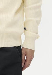 Tejelt, bordás, világoskrém pulóver hosszú ujjal. Szoros illeszkedésű a mandzsettáknál és oldalsó címkével rendelkezik. Fekete nadrággal viselve.