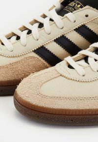 Ruskeat ja beige nubuk-nilkkurit mustilla nahkasuorilla, litteillä valkoisilla nauhoilla, kumipohjalla ja teksturoidulla kärjellä. Korostaa ommeltuja yksityiskohtia.