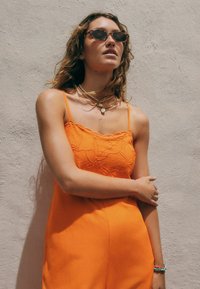 Robe sans manches orange avec broderies florales, texture lisse. Des colliers superposés et des lunettes de soleil complètent le look. Fond clair.