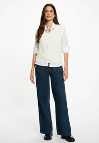 Gilet bianco lavorato a maglia con motivo a trecce e bottoni decorativi, abbinato a una camicia bianca con colletto e jeans a gamba larga blu scuro.