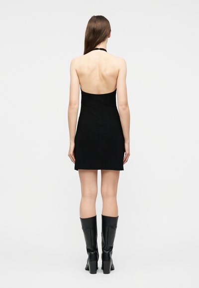 Γυναίκα που στέκεται, φοράει ένα μίνι μαύρο φόρεμα χωρίς μανίκια με halter και μαύρες μπότες με τακούνι έως το γόνατο, εμφανίζεται από πίσω σε απλό φόντο.