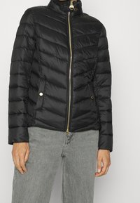Schwarze Daunenjacke mit hohem Kragen, goldenem Reißverschluss und horizontalen Nähten. Verfügt über seitliche Taschen mit goldenen Akzenten und eine taillierte Passform.