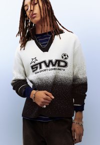 Pull en maille blanc dégradé vers le noir, avec un col en V, un logo graphique et des accents à pois. En dessous, une couche rayée bleu marine est visible.