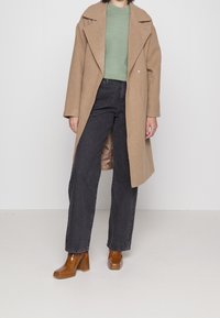 Mantel aus Kamelhaar-Mischung mit breitem Revers, kombiniert mit einem hellgrünen Pullover, schwarzen weiten Jeans und braunen Blockabsatzschuhen.