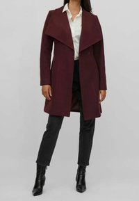 Abrigo de mezcla de lana burdeos con solapas anchas, mangas largas y diseño de apertura frontal, combinado con vaqueros negros y botas de punta.