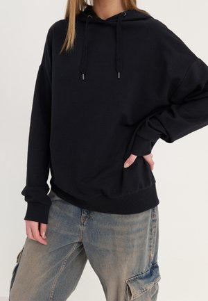 Osoba ubrana w czarną bluzę z kapturem i sznurkami oraz jasnoniebieskie luźne jeansy cargo, z ręką na biodrze, stojąca na neutralnym tle.