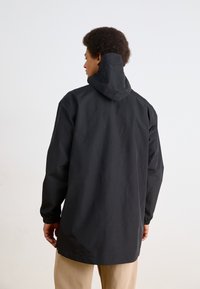 Zwarte lange jas met een capuchon, voorzien van een gladde textuur en elastische manchetten. Ontworpen met een iets losse pasvorm en een minimalistische stijl.