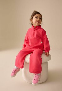Survêtement en polaire rose pour enfants avec col zippé, coupe oversize, poignets côtelés et poche avant. Associé à des baskets roses sur un tabouret blanc.