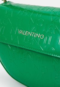 Grön, glansig handväska med präglad "VALENTINO"-logotyp, slät textur och en strukturerad form. Öppen stängning med guldfärgad hårdvara.