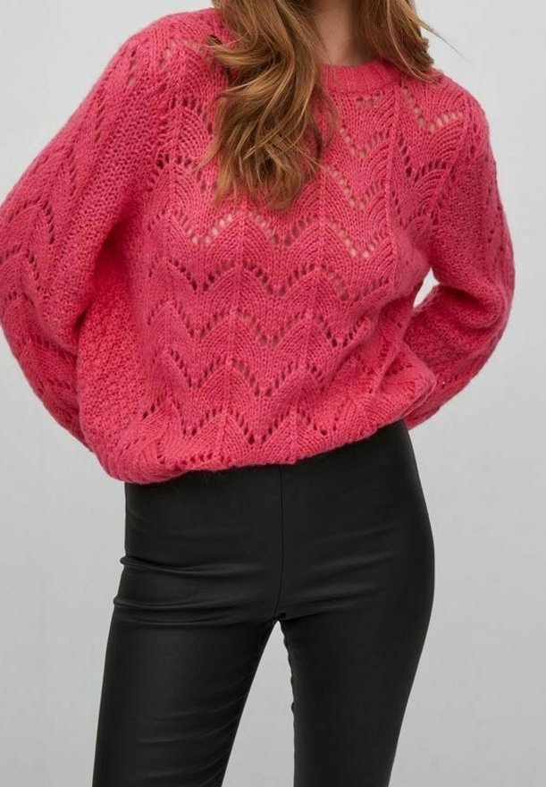 Femme portant un pull en maille épaisse rose vif avec un motif chevron et un pantalon en cuir noir taille haute, debout les mains dans le dos.