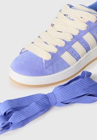 Purple suede sneaker with cream laces and three white stripes. Includes matching blue shoelaces beside the shoe. Flat rubber sole.