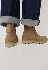 Bottines en suede marron avec élasthanne sur les côtés et fermeture éclair, portées avec un jean bleu foncé retroussé sur un fond de sol blanc.