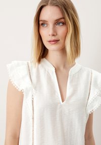 Blusa bianca leggera con maniche corte arricciate, bordatura in pizzo e scollo a V. Presenta strisce verticali lungo i bordi per una texture aggiuntiva.