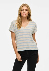 Top a righe con scollatura a V in beige e linee blu, maniche corte, vestibilità morbida, tessuto testurizzato, abbinato a pantaloni neri, sfondo neutro.