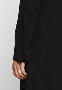 Veste noire sur mesure en tissu lisse, avec un bouton unique au poignet et une silhouette élégante et structurée. Main tenant la veste.