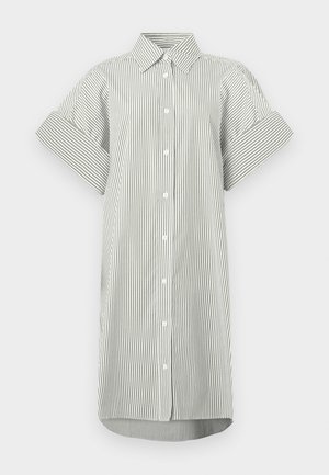 Femme portant une robe-chemise à manches courtes à rayures verticales, avec des bottes noires mi-mollet, tenant un sac à main noir sur un fond uni.
