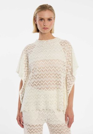 Femme portant un haut en dentelle blanche et un pantalon assorti en dentelle, debout devant un fond blanc uni, regardant droit devant elle.