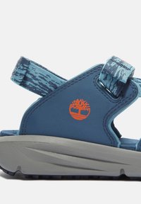 Siniset sandaalit, joissa on pehmeä, teksturoitu hihna ja kestävä pohja. Ikoninen oranssi logo sivulla. Säädettävät tarrakiinnitykset.
